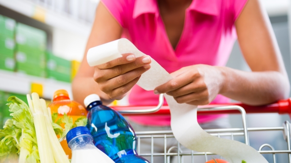 Unrecognizable woman checking a long grocery receipt leaning to a full shopping cart at store.