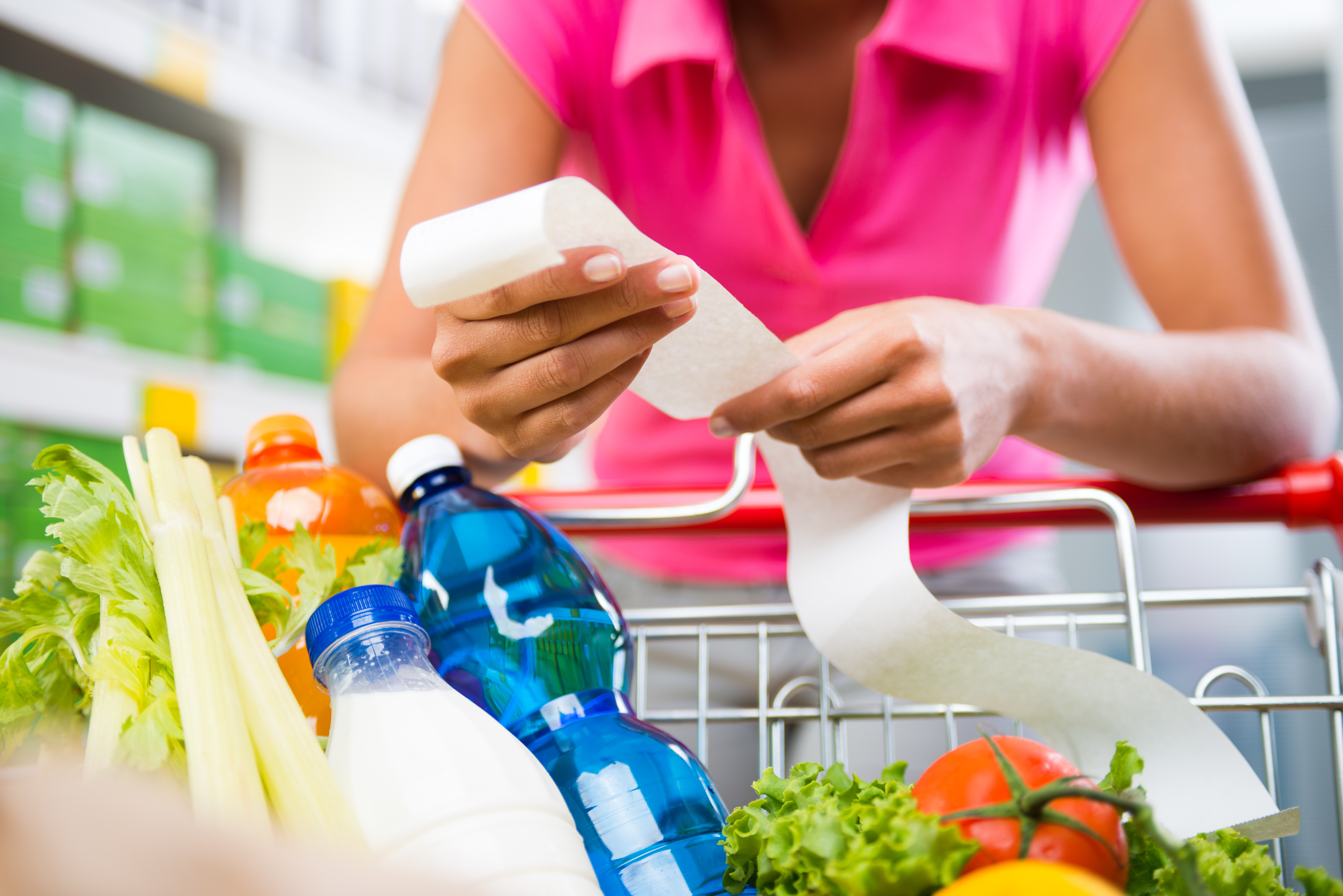 Unrecognizable woman checking a long grocery receipt leaning to a full shopping cart at store.