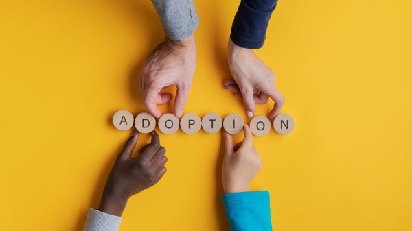 Hands of a family with black and white children assembling a word Adoption spelled on wooden cut circles. Over yellow background.