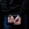 Teenage boy in black hoodie and jeans is arrested with hands cuffed behind his back. Dark studio background emphasizes his confinement. Represents crime, justice and consequence for youth.