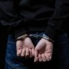 Teenage boy in black hoodie and jeans is arrested with hands cuffed behind his back. Dark studio background emphasizes his confinement. Represents crime, justice and consequence for youth.