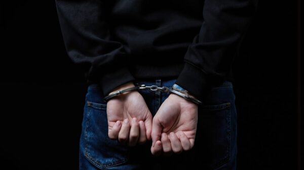 Teenage boy in black hoodie and jeans is arrested with hands cuffed behind his back. Dark studio background emphasizes his confinement. Represents crime, justice and consequence for youth.
